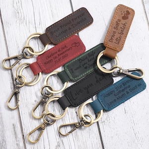 Op de afbeelding: Een verzameling gepersonaliseerde leren sleutelhangers in verschillende kleuren, waaronder bruin, rood, groen, zwart en blauw. Elke sleutelhanger heeft gegraveerde tekst met berichten zoals "Best Friend Forever" en "Drive Safe". Ze hebben goudkleurige sleutelringen en karabijnhaken.