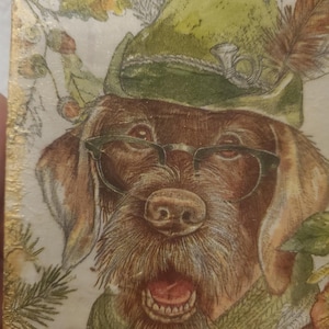 May include: A rectangular decorative tile featuring a brown dog wearing a green hat with a feather and a small horn emblem, along with black-rimmed glasses. The dog has a green scarf around its neck, and the edges of the tile are finished in gold.