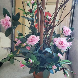 May include: A pink rose bush with multiple blooms in a terracotta pot. The bush is arranged with brown branches and is in front of a framed picture.