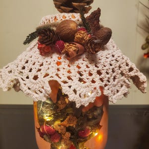 May include: A decorative, illuminated jar with a peach-colored exterior. The jar is adorned with a cream-colored crocheted cover and topped with pinecones, nuts, and dried flowers. The interior is lit with warm white fairy lights, creating a cozy, autumnal ambiance.