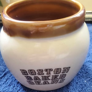 May include: A small, ceramic Boston Baked Beans pot with a white base and a brown rim. The words "BOSTON BAKED BEANS" are printed in a dark font on the front of the pot. The pot has a rounded shape.