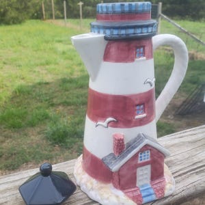 May include: A ceramic lighthouse-shaped pitcher featuring horizontal red and white stripes, a blue and black top, and a small attached house at the base. The pitcher has a white handle, a spout, and a separate black lid with a round knob handle.