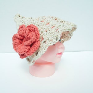 May include: A crocheted hat in a light beige colour, adorned with a large coral-coloured flower. The hat has an open weave design and is shown on a mannequin head. The flower is prominently displayed on the side of the hat.
