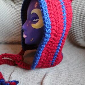 May include: A handmade crochet hood in red with blue trim and accents. The hood is displayed on a purple mannequin head with a yellow crescent moon design on the face. The hood has long ties.