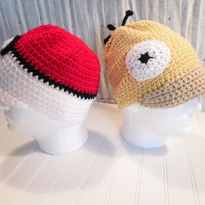 K&ouml;nnte beinhalten: Zwei geh&auml;kelte M&uuml;tzen, eine rot, wei&szlig; und schwarz mit einem Pokeball-Design, und die andere gelb, wei&szlig; und braun mit einem Cartoon-Augen-Design.