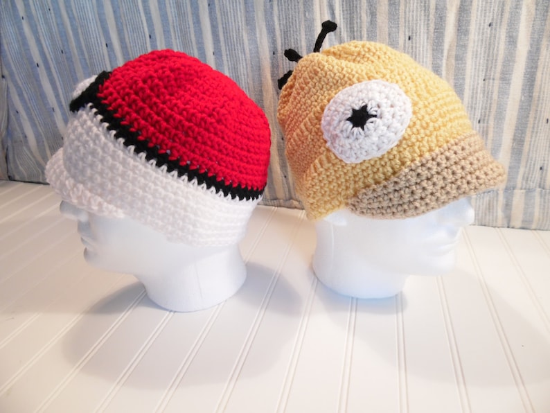 K&ouml;nnte beinhalten: Zwei geh&auml;kelte M&uuml;tzen, eine rot, wei&szlig; und schwarz mit einem Pokeball-Design, und die andere gelb, wei&szlig; und braun mit einem Cartoon-Augen-Design.