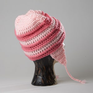 May include: Pink and light pink crocheted hat with a long tail. The hat is on a wooden display stand.