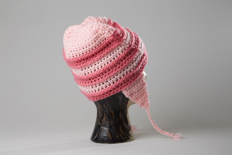 May include: Pink and light pink crocheted hat with a long tail. The hat is on a wooden display stand.