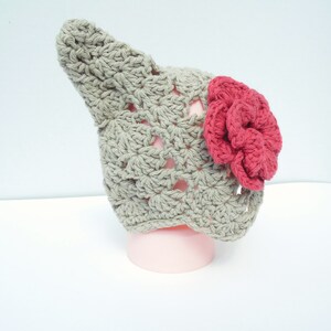 Op de afbeelding: Een handgemaakte gehaakte hoed in een lichtgrijze kleur, met een grote, levendige rode bloem aan de zijkant. De hoed heeft een uniek, puntig ontwerp, perfect voor een baby of kind. De hoed wordt getoond op een roze vorm.