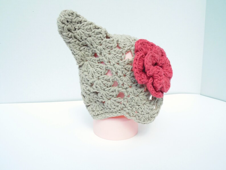 Op de afbeelding: Een handgemaakte gehaakte hoed in een lichtgrijze kleur, met een grote, levendige rode bloem aan de zijkant. De hoed heeft een uniek, puntig ontwerp, perfect voor een baby of kind. De hoed wordt getoond op een roze vorm.