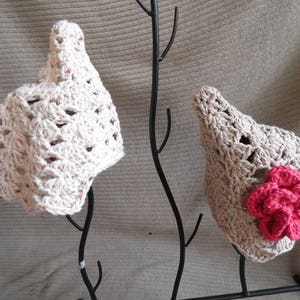 Op de afbeelding: Twee gehaakte hoeden. E&eacute;n is gebroken wit met roze spikkels, de andere is beige met een grote roze bloem. Beide hoeden hebben een puntige kroon en een open weefpatroon. Ze worden tentoongesteld op een zwarte metalen standaard.