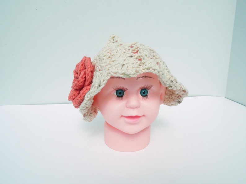May include: A cream-coloured, crocheted sun hat with a wide brim and a large coral-coloured flower on the side. The hat is displayed on a pink mannequin head with blue eyes.