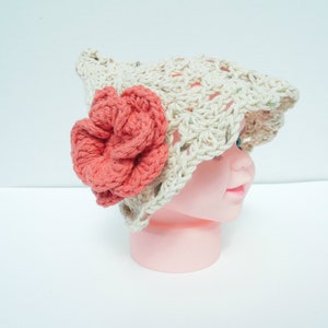May include: A crocheted hat in a light beige colour, adorned with a large coral-coloured flower. The hat has an open weave design and a brim. This handmade hat is a stylish accessory for babies and children.