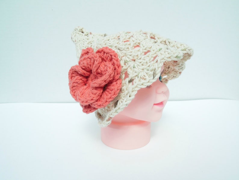 May include: A crocheted hat in a light beige colour, adorned with a large coral-coloured flower. The hat has an open weave design and a brim. This handmade hat is a stylish accessory for babies and children.