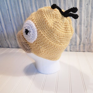 K&ouml;nnte beinhalten: Eine gelbe H&auml;kelm&uuml;tze mit einer braunen Nase, einem wei&szlig;en Auge mit einem schwarzen Stern und schwarzen Antennen. Die M&uuml;tze sitzt auf einem wei&szlig;en Mannequin-Kopf.