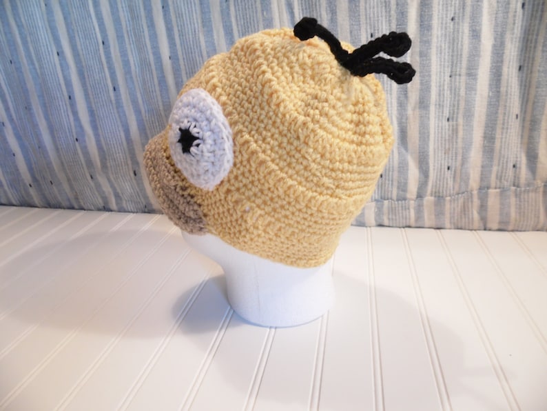 K&ouml;nnte beinhalten: Eine gelbe H&auml;kelm&uuml;tze mit einer braunen Nase, einem wei&szlig;en Auge mit einem schwarzen Stern und schwarzen Antennen. Die M&uuml;tze sitzt auf einem wei&szlig;en Mannequin-Kopf.