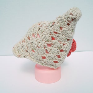 May include: A light beige crocheted hat with a floral pattern and a small red accent. The hat is displayed on a pink cylinder. The crochet work is visible, showcasing the texture and design.