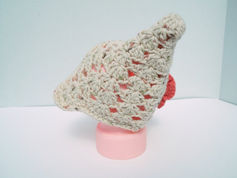 May include: A light beige crocheted hat with a floral pattern and a small red accent. The hat is displayed on a pink cylinder. The crochet work is visible, showcasing the texture and design.