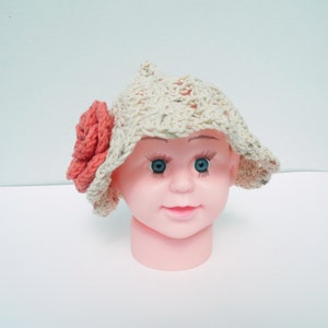 May include: A cream-coloured, crocheted hat with a wide brim and a large coral-coloured flower on the side. The hat is displayed on a pink mannequin head with blue eyes.