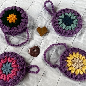 May include: Four handmade crochet coasters in shades of purple, with colorful floral centers. Two small heart-shaped wooden embellishments are also present. The coasters have a circular shape with a loop for hanging.