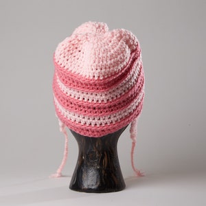 May include: A pink and white striped crocheted hat with long ties. The hat is displayed on a wooden mannequin.