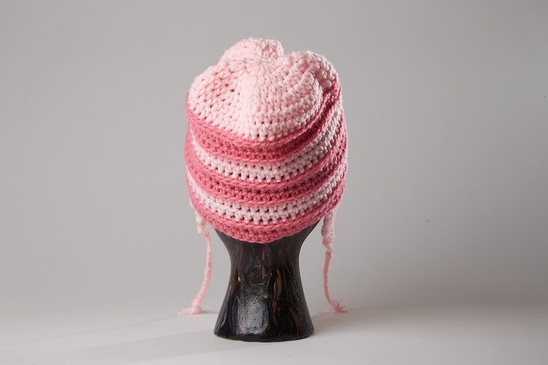 May include: A pink and white striped crocheted hat with long ties. The hat is displayed on a wooden mannequin.
