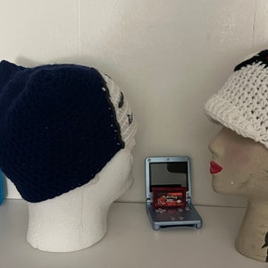 K&ouml;nnte beinhalten: Zwei geh&auml;kelte M&uuml;tzen, eine blaue und eine wei&szlig;e mit roten und schwarzen Streifen, werden auf Mannequin-K&ouml;pfen pr&auml;sentiert. Eine blaue Game Boy Advance SP mit einer roten Pokemon-Spielkassette befindet sich in der Mitte des Bildes.