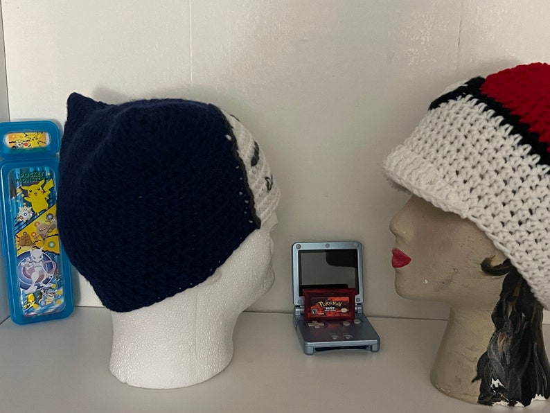 K&ouml;nnte beinhalten: Zwei geh&auml;kelte M&uuml;tzen, eine blaue und eine wei&szlig;e mit roten und schwarzen Streifen, werden auf Mannequin-K&ouml;pfen pr&auml;sentiert. Eine blaue Game Boy Advance SP mit einer roten Pokemon-Spielkassette befindet sich in der Mitte des Bildes.