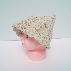 May include: A light beige crocheted hat with a flared brim and a pointed crown. The hat features an open weave design with green and brown flecks. It is displayed on a pink mannequin head, showcasing the hat's shape and texture.