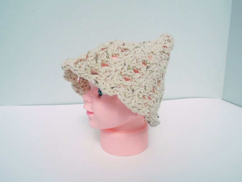 May include: A light beige crocheted hat with a flared brim and a pointed crown. The hat features an open weave design with green and brown flecks. It is displayed on a pink mannequin head, showcasing the hat's shape and texture.