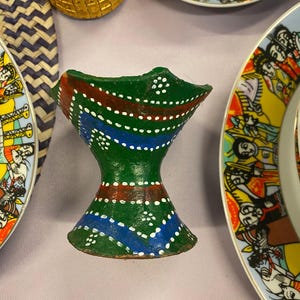 Könnte beinhalten: Ein handbemalter, grüner Keramik-Räuchergefäß mit roten und blauen Akzenten und weißen Punktdetails. Der Brenner hat eine ausgestellte Ober- und Unterseite und ist von bunten Tellern und einem goldenen Behälter umgeben.