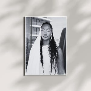 May include: Black and white portrait print of a person wearing a white dress and veil, with long, wavy hair and dangling earrings. The background includes a building and a draped fabric.