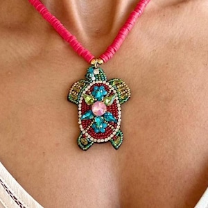 Puede incluir: Collar con colgante de tortuga con cuentas y cordón rosa intenso. El colgante de tortuga presenta un centro rojo con piedras preciosas azules, verdes y rosas, y está delineado con cuentas verdes y doradas. Un accesorio vibrante.