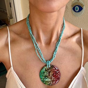 May include: A turquoise beaded necklace with a large, round pendant. The pendant features a colorful design with green, red, and white accents. The necklace is worn with a cream-colored top and gold earrings.