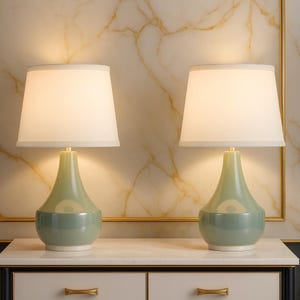 May include: Two matching table lamps with off-white shades and light green bases. The lamps have a classic, elegant design, with a brass-colored neck and a rounded base. The lamps are placed on a white marble-topped cabinet.