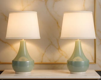May include: Two matching table lamps with off-white shades and light green bases. The lamps have a classic, elegant design, with a brass-colored neck and a rounded base. The lamps are placed on a white marble-topped cabinet.