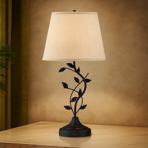 May include: A black metal table lamp with a beige fabric shade. The lamp base features a decorative vine design with leaves. The lamp is on a wooden surface.