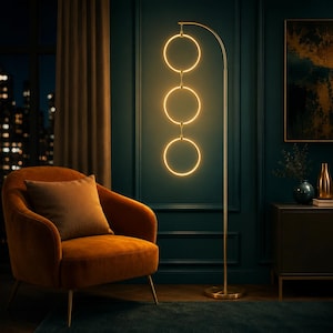 May include: A modern gold floor lamp with three illuminated circular lights. The lamp is in a room with an orange armchair, a brown pillow, and a dark teal wall. The room has a dark blue rug and a window with a city view.