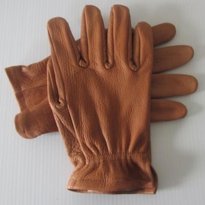 May include: A pair of brown leather gloves with a textured surface. The gloves are folded in half, with the fingertips of the left hand visible.