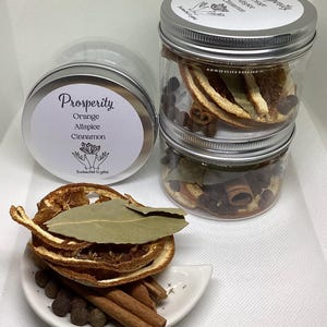 May include: Three clear jars with silver lids, stacked and filled with dried orange slices, cinnamon sticks, and allspice berries. One jar is separate, and another jar is on its side. The jars have labels that read "Prosperity" with ingredient names.
