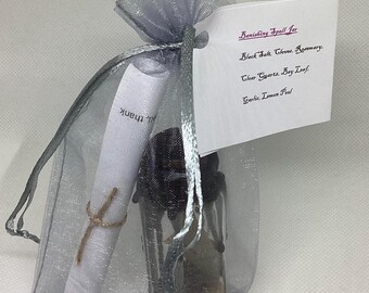 May include: A small, clear glass jar filled with dark contents, a rolled scroll, and a tag with handwritten text, all inside a sheer silver drawstring bag. The tag reads "Banishing Spell Jar" with a list of ingredients.