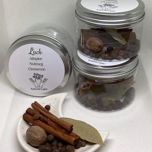 May include: Three clear jars with silver lids, stacked, filled with spices. A small white dish holds cinnamon sticks, nutmeg, peppercorns, and a bay leaf. The jars and dish sit on a white surface. Text on the jars reads "Luck, Allspice, Nutmeg, Cinnamon."