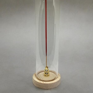 May include: A clear glass incense holder with a wooden base. A red incense stick is centered inside the glass. The base is a light-colored wood with a circular shape. The holder has a gold-colored metal incense stick holder.