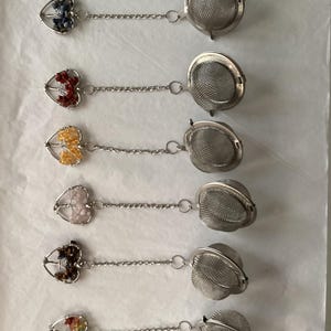 May include: Six tea infusers with heart-shaped gemstone accents and stainless steel mesh infuser baskets. Each infuser has a different gemstone color, including blue, red, yellow, pink, brown, and a rainbow of colors.