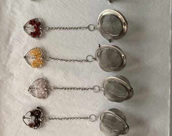 Op de afbeelding: Zes thee-infusers met hartvormige edelsteenaccenten en roestvrijstalen mesh-infuserkorven. Elke infuser heeft een andere edelsteentint, waaronder blauw, rood, geel, roze, bruin en een regenboog.
