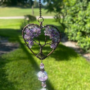 May include: A heart-shaped suncatcher with a tree design, crafted from copper wire and adorned with amethyst crystals. A clear crystal ball with the words "Made with Love" hangs below. The suncatcher is suspended by a chain.