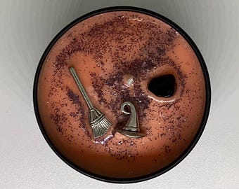 May include: A round, orange-colored candle with a black rim, decorated with silver charms of a witch's hat and broom. The candle is sprinkled with purple glitter and has a black stone accent.