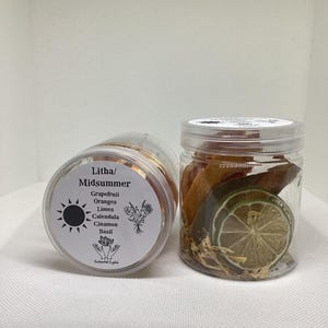 May include: Two clear jars filled with dried citrus slices and herbs. One jar is tilted, revealing the contents and a label with the text "Litha/Midsummer" and ingredient names. The other jar is upright, showcasing the contents.