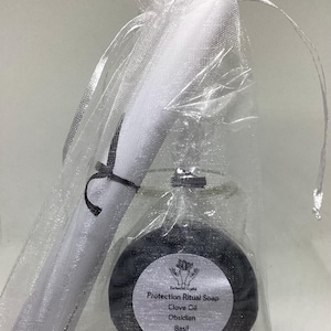 May include: A gift set in a sheer white drawstring bag. The set includes a white scroll tied with a black ribbon and a round, dark gray soap with a label that reads "Protection Ritual Soap, Clove Oil, Obsidian, Basil."