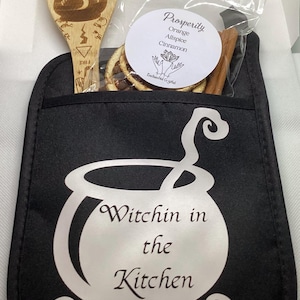 May include: A black pot holder with the text "Witchin in the Kitchen" in white. It includes a wooden spoon with etched designs and a bag of spices labeled "Prosperity". The bag contains orange slices and cinnamon sticks.